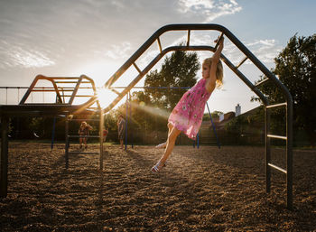 Side view of girl hanging on monkey bars against sky at playground during sunset