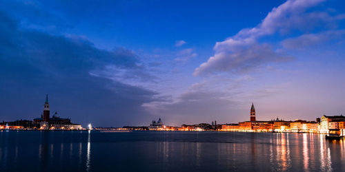 Illuminated buildings by river against cloudy sky