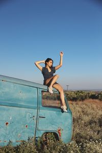 Young woman with arms raised sitting on abandoned van against clear sky