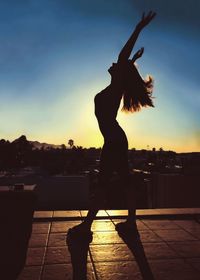 Silhouette of woman with arms raised standing on building terrace during sunset