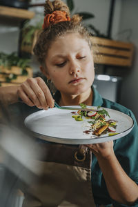 Young chef arranging vegetable with tongs in plate while standing in kitchen