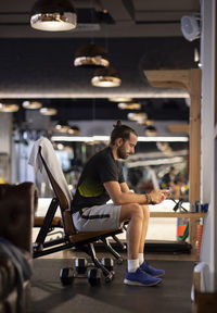 Side view of adult sportsman sitting on bench and browsing smartphone during break in contemporary gym