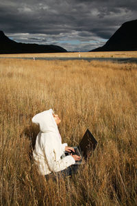 Mid adult woman using laptop while sitting on grassy field against cloudy sky