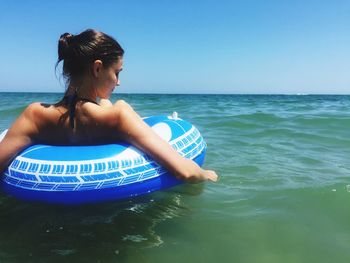 Rear view of young woman with inflatable ring floating in sea against clear blue sky during sunny day