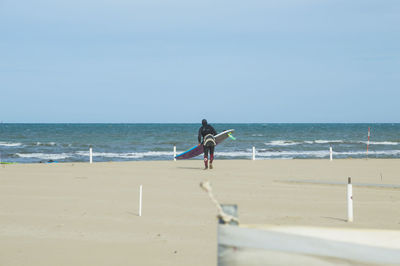 Rear view of person with surfboard walking at beach towards sea against clear sky