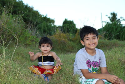 Portrait of cute siblings sitting on grass against sky