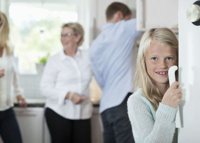 Portrait of smiling girl holding door handle with family in background at kitchen