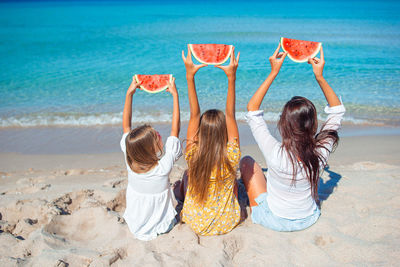 Rear view of mother and daughters holding watermelon sitting on beach