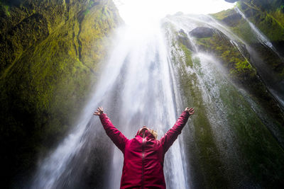 Low angle view of mid adult woman with arms raised standing against waterfall