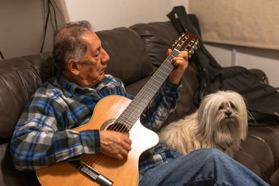 Senior man playing guitar while sitting on sofa with dog at home