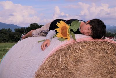 Boy with sunflower lying on hay bale against sky at farm