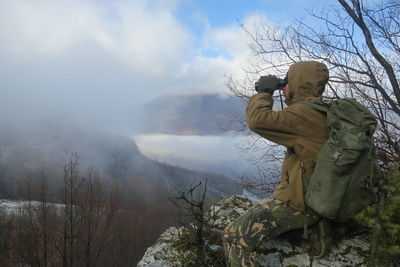 Side view of army soldier using binoculars while sitting on cliff against sky during foggy weather