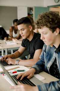 Male students using laptop while sitting by table in classroom