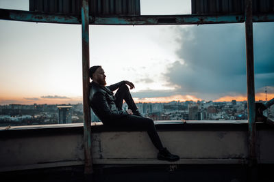 Side view of man sitting on window at sunset