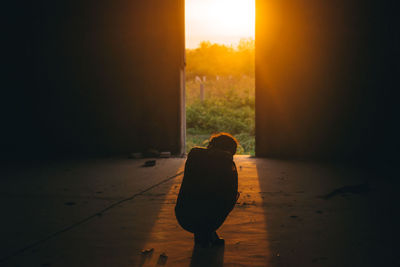 Rear view of silhouette girl crouching in darkroom during sunset