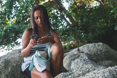 Young woman using phone while sitting on rock