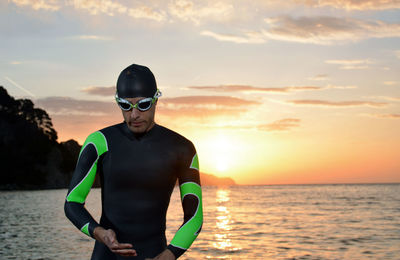 Man wearing wetsuit while standing at beach against orange sky