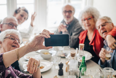 Woman clicking photograph of senior friends through smart phone while sitting in restaurant