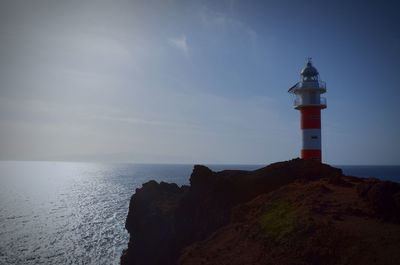 Lighthouse on cliff by sea against sky