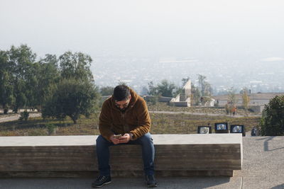 Young man sitting on retaining wall against sky