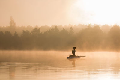 Silhouette woman meditating in lake against sky during sunrise