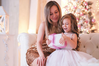 A fasion woman is presenting christmas gift for daughter sitting on the sofa in light living room.