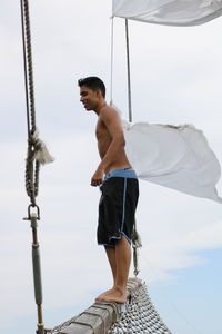 Side view of shirtless man standing on boat against cloudy sky