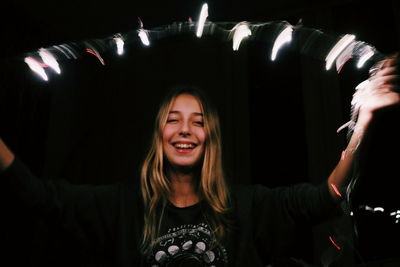 Close-up of young woman holding illuminated string light while standing against black background