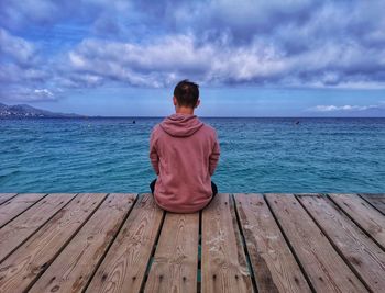 Rear view of man sitting on pier over sea against sky