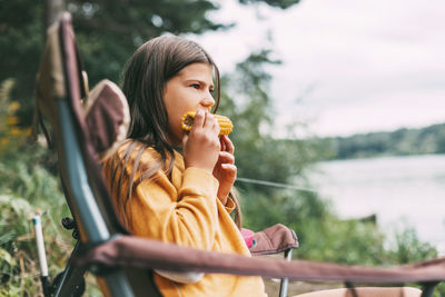 A teenage girl in a bright yellow sweater is sitting in a camping chair on the shore of the lake 