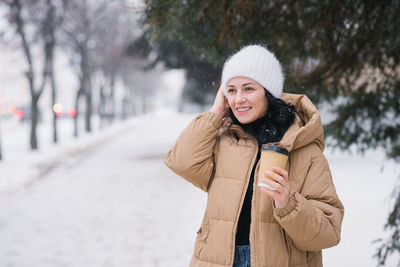 A cute girl smiles and holds a disposable cup of coffee. adjust the hat in winter under the snow.