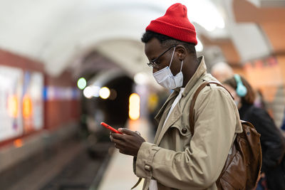 African man in face mask in subway station wait for train, using smartphone, scrolling social media