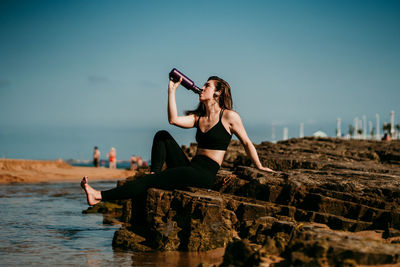 Peaceful female in sportswear sitting on stone near river drinking water while doing yoga