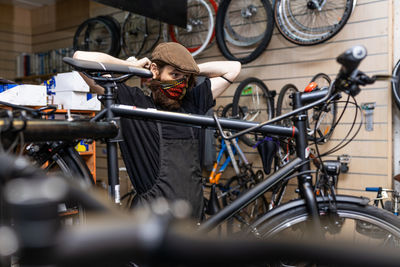 Serious bearded male master in workwear putting on protective mask while working in bicycle service workshop during coronavirus pandemic