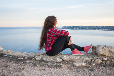 Woman sitting on rock looking at sea against sky