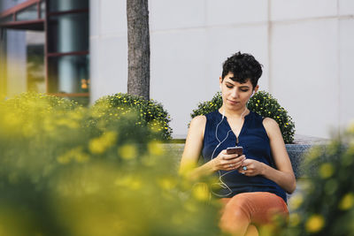 Woman listening music while sitting on retaining wall against building