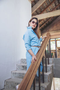 Portrait of young woman standing on staircase against wall