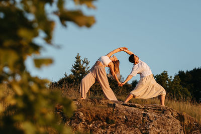 Couple making beautiful pose, man and woman practicing yoga outdoors on the rocky hill at sunset