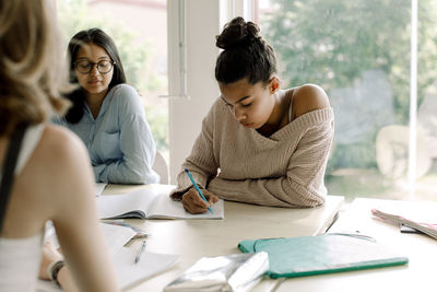 Teenage student writing while sitting by table against glass window in classroom