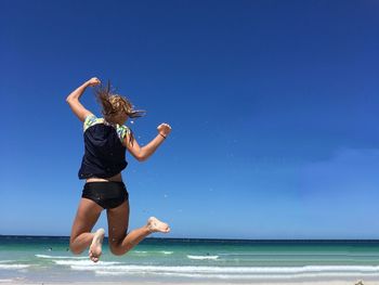 Rear view of woman jumping at beach against blue sky