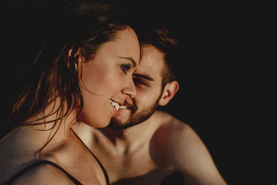 Smiling young couple in swimwear and with wet hair cuddling while resting on dark beach at sunset together during romantic holidays