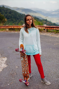 Woman holding skateboard while standing on road against mountains