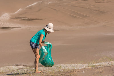 Mature woman in a white hat and green gloves collecting garbage in a green bag. concept plogging