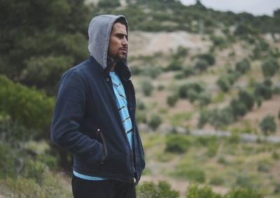 Thoughtful young man wearing hooded shirt looking away while standing on hill