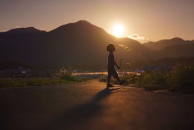 Silhouette boy walking on road towards mountain against sky during sunset