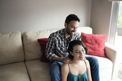 Smiling boyfriend massaging girlfriend's neck while sitting on sofa at home