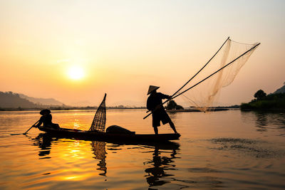 Silhouette of people fishing in boat on lake against sky during sunset