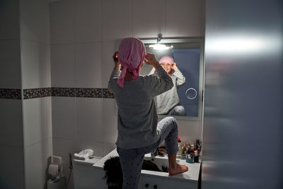 Back view of sick little child putting on pink bandana in front of mirror in bathroom