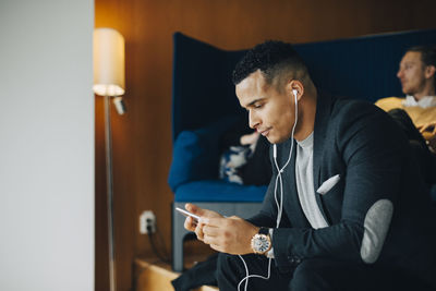 Mid adult businessman listening to in-ear headphones while using smart phone at office cafeteria