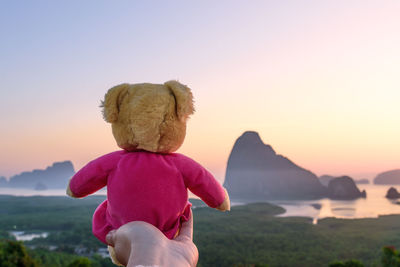 Cropped hand holding teddy bear against sea during sunset